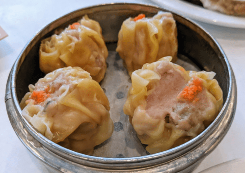 A metal steamer basket containing four pieces of siu mai, a type of dumpling, topped with orange fish roe.