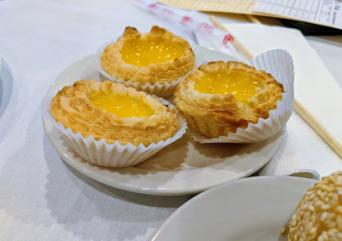 Three golden egg tarts in paper cups on a white plate, resting on a table with a napkin and chopsticks nearby.