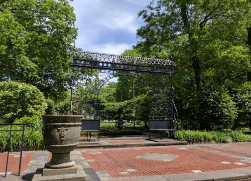 A decorative metal archway surrounded by lush greenery and a stone planter on a brick pathway.