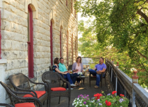 A group of four people relax on a balcony surrounded by greenery, enjoying drinks and conversation.