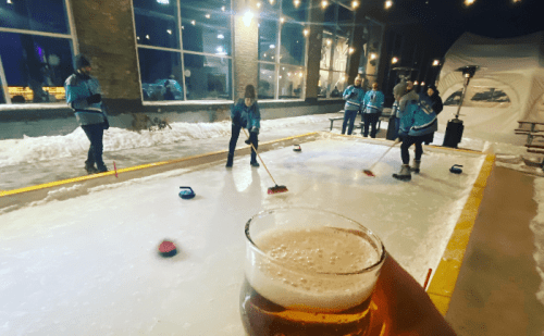 A person holds a beer while others curl on an outdoor ice rink, surrounded by snow and string lights.