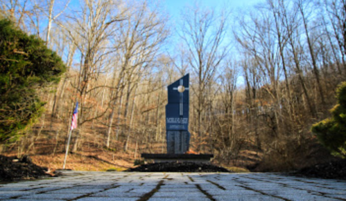 A tall monument stands in a wooded area, with a flag nearby and trees in the background under a clear blue sky.