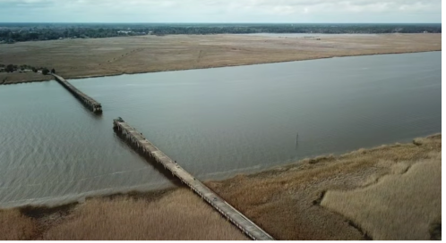 Aerial view of a long pier extending into a calm body of water, surrounded by marshland and cloudy skies.