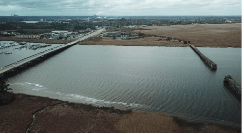 Aerial view of a river with a bridge, marshland, and industrial area in the background under a cloudy sky.