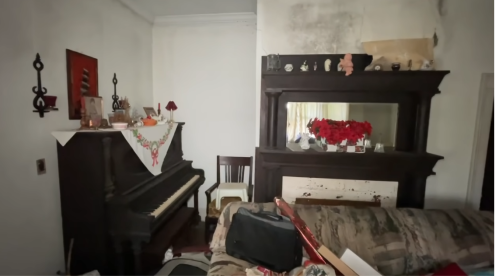 A cluttered living room featuring a piano, a mirror, and decorative items on a mantel, with a couch in the foreground.