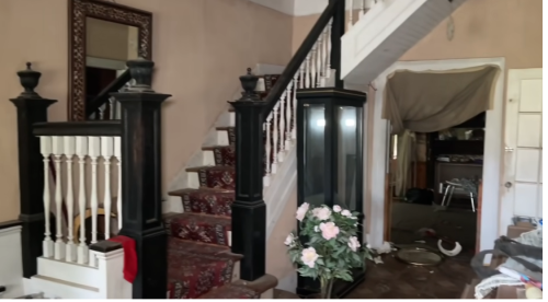 A staircase with a patterned carpet, a mirror, and a glass display case, leading to a cluttered room.