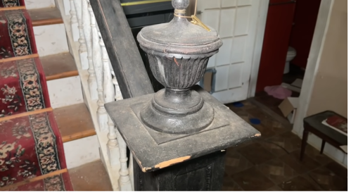 A close-up of a dusty, ornate stair banister post with a decorative finial, set against a staircase and cluttered background.