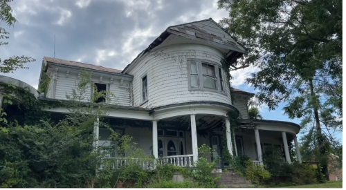An old, dilapidated Victorian house with overgrown vegetation and a cloudy sky in the background.