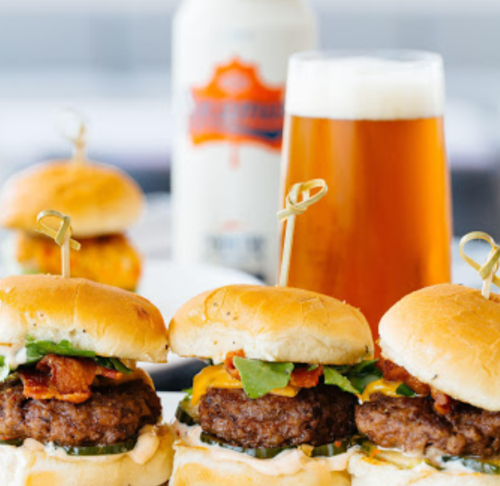 Three mini burgers with bacon, cheese, and lettuce, served with a pint of beer in the background.
