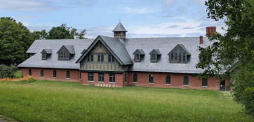 A large, historic brick building with a gray roof, surrounded by green grass and trees under a cloudy sky.