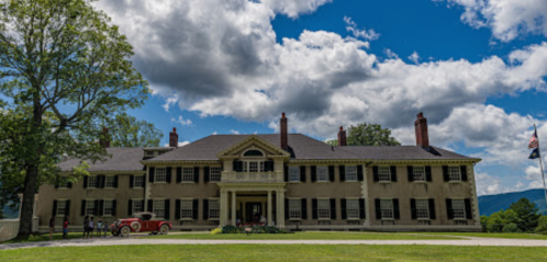 A large historic mansion with a green lawn, surrounded by trees and under a blue sky with fluffy clouds.