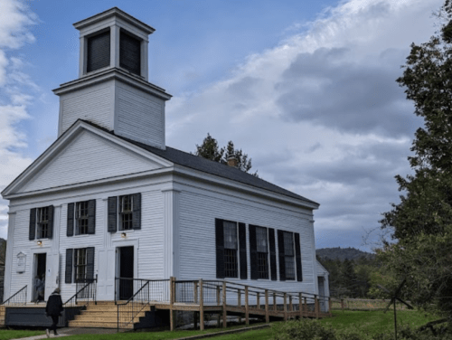 A white historic building with a bell tower, surrounded by greenery and under a cloudy sky.