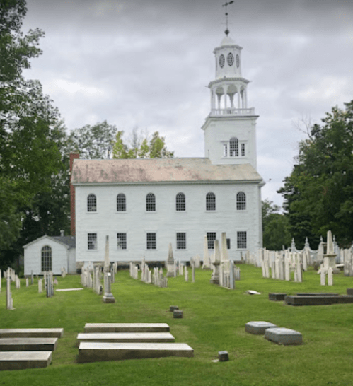 A historic white church stands behind a cemetery filled with gravestones, under a cloudy sky.