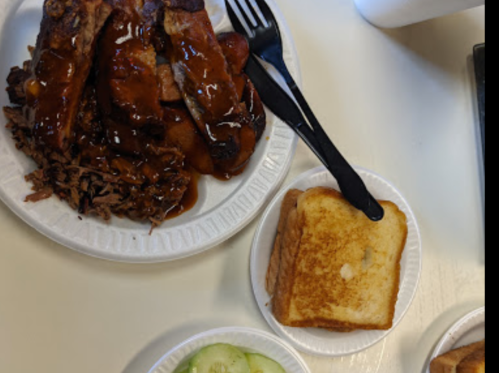 A plate with barbecue ribs and pulled pork, accompanied by a slice of toasted bread and a small salad of cucumber slices.