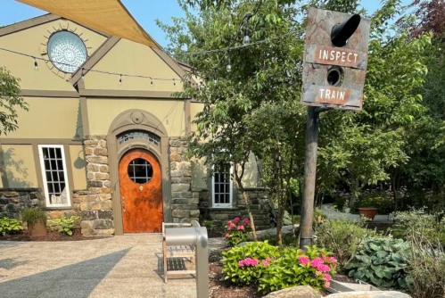 A rustic building with a round window and wooden door, next to a weathered sign reading "INSPECT TRAIN" amidst greenery.