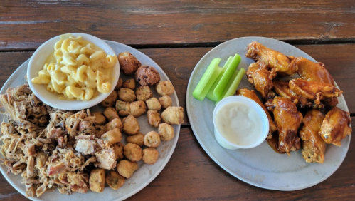 A plate of pulled pork, fried okra, and macaroni, alongside a plate of buffalo wings with celery and ranch dressing.