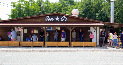A rustic wooden building with a sign reading "Jon G's," featuring people waiting outside on a sunny day.
