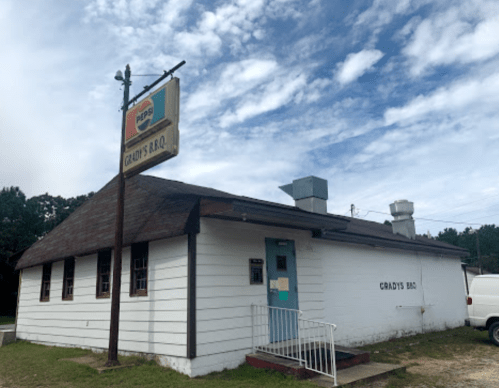 A small, white building with a brown roof and a sign reading "Grady's B.Q." under a cloudy sky.