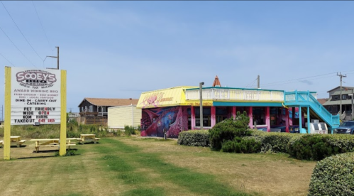 Colorful restaurant with outdoor seating and a sign, surrounded by grass and shrubs, in a coastal setting.