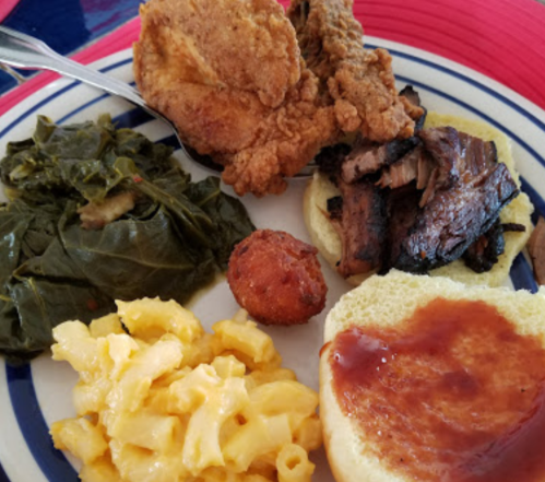A plate of Southern food featuring fried chicken, collard greens, macaroni and cheese, cornbread, and a bun with sauce.