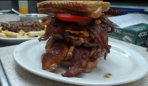 A towering burger with multiple beef patties, bacon, cheese, tomato, and pickles, served on toasted bread.