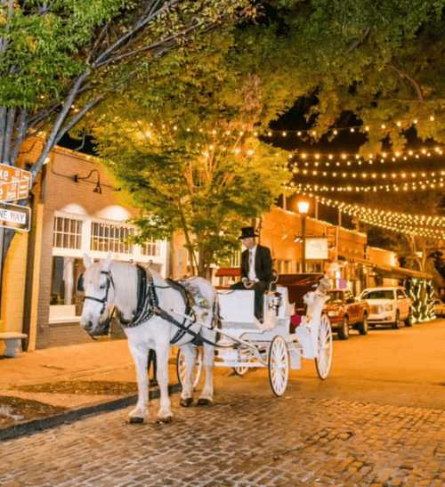 A horse-drawn carriage waits on a cobblestone street, illuminated by string lights and surrounded by vintage cars.