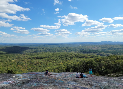 Three people sit on a rocky ledge, overlooking a vast green landscape under a blue sky with fluffy clouds.