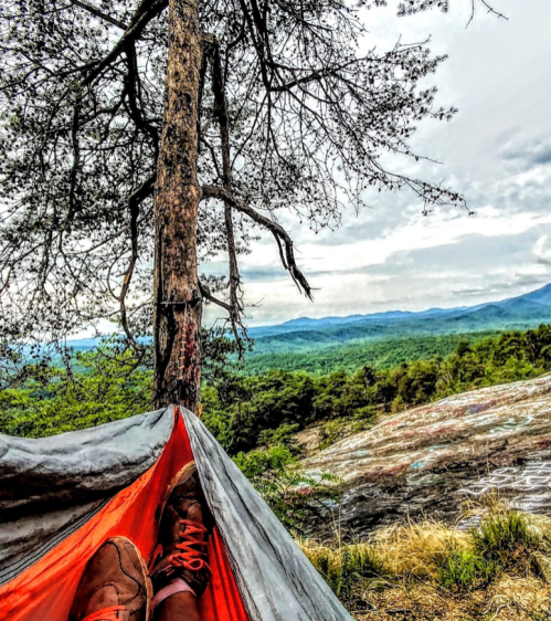 A view from a hammock with feet visible, surrounded by trees and mountains under a cloudy sky.