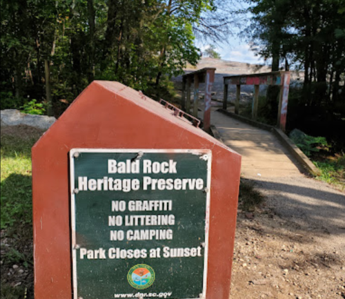 Sign at Bald Rock Heritage Preserve with rules: no graffiti, no littering, no camping; bridge in the background.