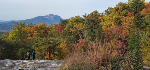 Two hikers walk along a rocky path surrounded by vibrant autumn foliage and distant mountains.
