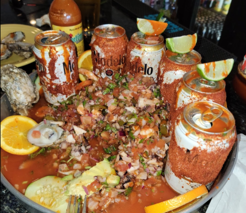 A large platter of seafood cocktail with cans of Modelo beer, garnished with lime, orange, and spices.