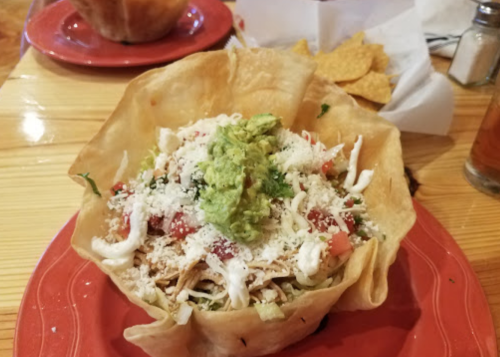 A colorful taco salad in a crispy tortilla bowl, topped with guacamole, cheese, tomatoes, and sour cream.