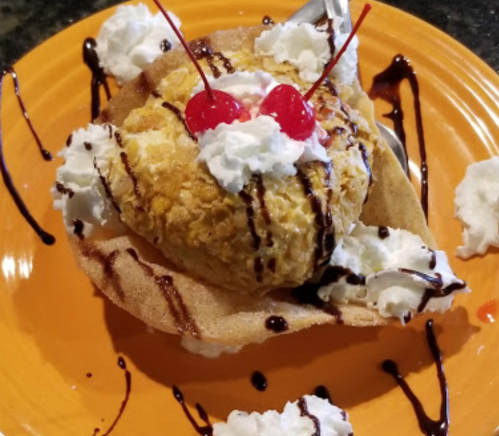 A dessert featuring a large scoop of ice cream in a waffle bowl, topped with whipped cream and cherries, drizzled with chocolate.