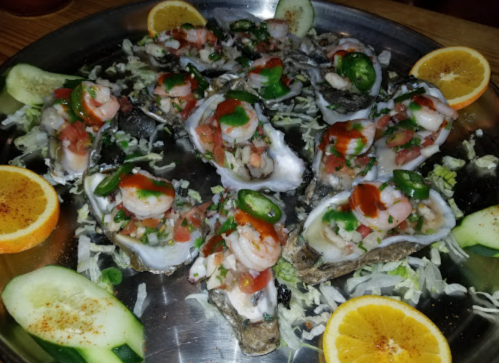 A platter of oysters topped with shrimp, salsa, jalapeños, and garnished with cucumber and orange slices.