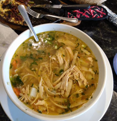 A bowl of chicken soup with vegetables and herbs, served with a spoon on a table.