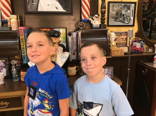 Two young boys with short haircuts smile in a vintage barbershop, surrounded by memorabilia and decor.