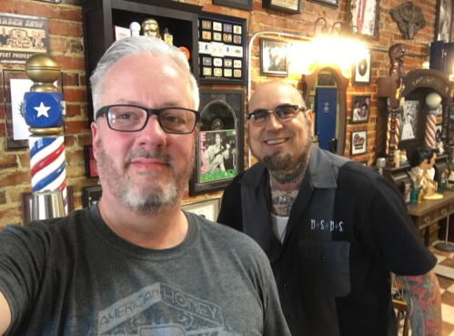 Two men smiling for a selfie in a vintage barbershop filled with memorabilia and decor.