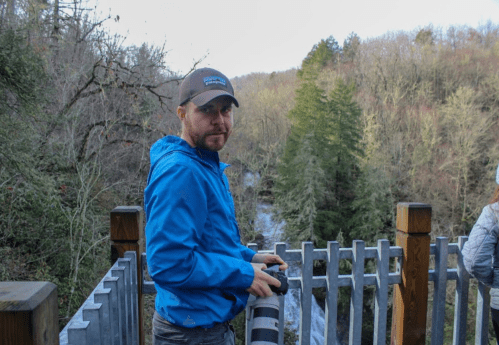 A man in a blue jacket stands on a lookout, holding a camera, with a waterfall and trees in the background.