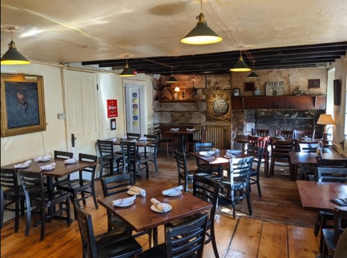 Cozy restaurant interior with wooden tables, black chairs, and a stone fireplace, featuring warm lighting and artwork.