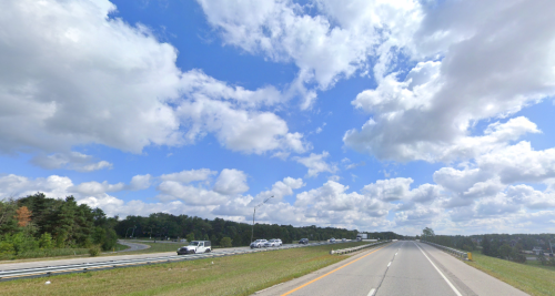 A wide highway under a blue sky filled with fluffy clouds, flanked by green trees on the left.