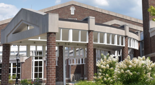 A modern building with large windows and a brick facade, surrounded by greenery and flowers under a blue sky.