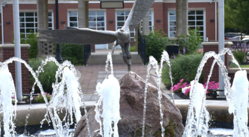 A bronze eagle sculpture perched on a rock, surrounded by water fountains in a landscaped area.