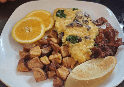 A plate with an omelette, bacon, seasoned potatoes, orange slices, and a piece of bread.