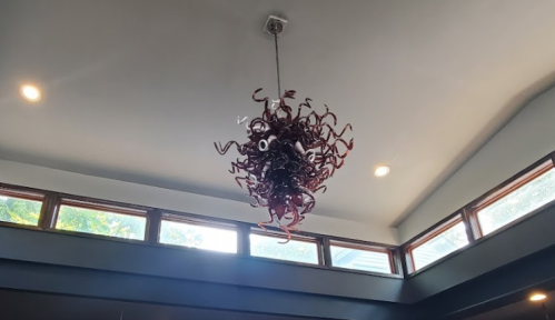 A unique, artistic chandelier with swirling red and brown shapes hangs from a ceiling with large windows.