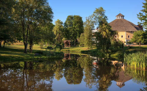 A serene landscape featuring a pond reflecting trees and a round building with a thatched roof under a clear blue sky.