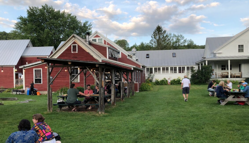 A rustic outdoor gathering space with people seated at picnic tables, surrounded by green grass and red barn-style buildings.