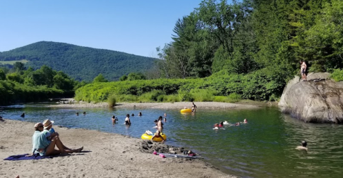 A sunny river scene with people swimming, lounging on the shore, and a rocky area surrounded by greenery and mountains.