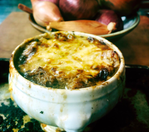 A bowl of French onion soup topped with melted cheese, with onions and a wooden spoon in the background.