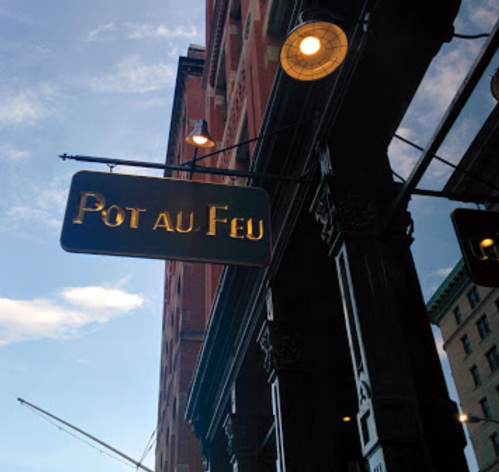 Sign for "Pot au Feu" hanging from a building, with a blue sky and clouds in the background.