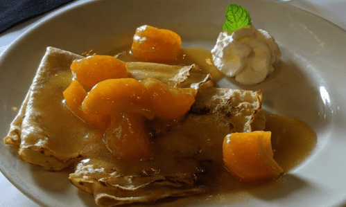 A plate of crepes topped with peach slices in syrup and a dollop of whipped cream, garnished with a mint leaf.